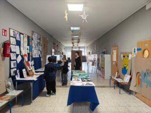 Szkolny korytarz zaaranżowany na galerię sztuki, w którym uczniowie oglądają liczne prace plastyczne wyeksponowane na stołach z niebieskimi obrusami, tablicach ściennych oraz szafkach.