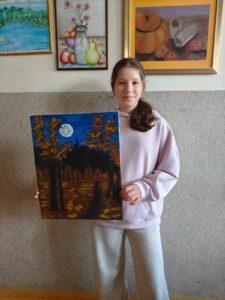 Uczennica swój obraz przedstawiający nocny las z księżycem w pełni, stojąc na tle ściany z innymi oprawionymi pracami plastycznymi
