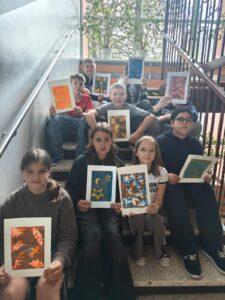 Grupa uczniów siedzi na szkolnych schodach, prezentując wykonane przez siebie różnorodne, kolorowe grafiki i prace plastyczne.