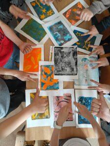 Widok z góry na stół, na którym leżą różnorodne grafiki wykonane przez uczniów, otoczone dłońmi autorów pokazujących gesty kciuka w górę i znaki zwycięstwa.