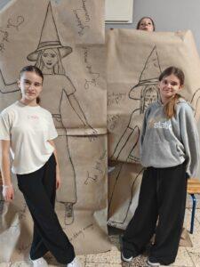 Dziewczyny stoją przed dużymi rysunkami postaci w kapeluszach czarownic