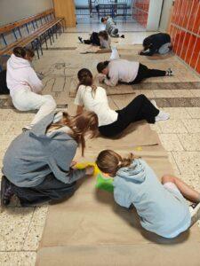 Uczniowie pracują wspólnie na podłodze szkolnego korytarza, tworząc kolorowe rysunki i napisy na dużych arkuszach brązowego papieru.