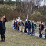 Kobieta w wianku na głowie prowadzi zajęcia terenowe dla grupy dzieci stojących w rzędzie na ścieżce, na tle lasu i zbocza kamieniołomu.