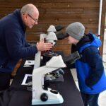 Mężczyzna pomaga chłopcu spojrzeć przez mikroskop ustawiony na stole w plenerze.