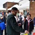 Mężczyzna w otoczeniu grupy dzieci i dorosłych podczas plenerowego wydarzenia edukacyjnego.