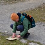 Chłopiec w pomarańczowej czapce i zielonej kurtce kuca na żwirowej ścieżce, uważnie oglądając mapę rozłożoną na trawiastym poboczu.