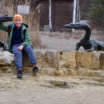 Chłopiec w pomarańczowej czapce siedzi uśmiechnięty na kamiennym murku, opierając rękę o rzeźbę prehistorycznego gada, na tle suchych traw i krzewów.