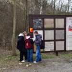 Trzy dziewczynki z plecakami stoją przy dużej tablicy edukacyjnej „Skały osadowe”, na której prezentowane są różne rodzaje minerałów i schematy geologiczne w terenie leśnym.