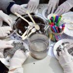 Dłonie w białych ochronnych rękawiczkach pracują przy okrągłym stole, mieszając ziemię i sypkie substancje na papierowych talerzach podczas warsztatów przyrodniczych.