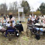 Grupa dzieci siedzących przy stolikach na zewnątrz unosi dłonie w białych rękawiczkach w geście radości podczas zajęć edukacyjnych w plenerze.