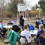 Kobieta w wianku prowadzi prelekcję dla dzieci siedzących przy okrągłych stolikach w plenerze, na tle białej budki meteorologicznej i wiosennej roślinności.