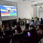 Grupa uczniów bierze udział w interaktywnym quizie o dinozaurach, korzystając ze smartfonów i patrząc na ekran projekcyjny z widocznym pytaniem oraz grafiką stegozaura.