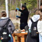 Mężczyzna w niebieskich rękawiczkach prezentuje uczniom doświadczenie chemiczne z użyciem kolb z kolorowymi płynami przy drewnianym stole w plenerze.