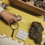 Widok z góry na stolik warsztatowy, przy którym uczestnicy badają minerały za pomocą lupy i przyrządów geologicznych.