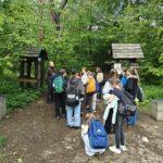 Grupa wycieczkowa z plecakami słucha przewodnika przed drewnianą bramą wejściową z napisem „Park Gródek”, otoczoną gęstą zielenią lasu.
