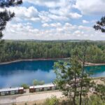Widok z góry na lazurowy zalew otoczony gęstym lasem liściastym, sfotografowany w słoneczny dzień przez ramę z gałęzi drzew iglastych.